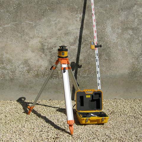 laser level on a gravel pathway with a stone wall behind it. The other elements of the laser level kit stand next to it.